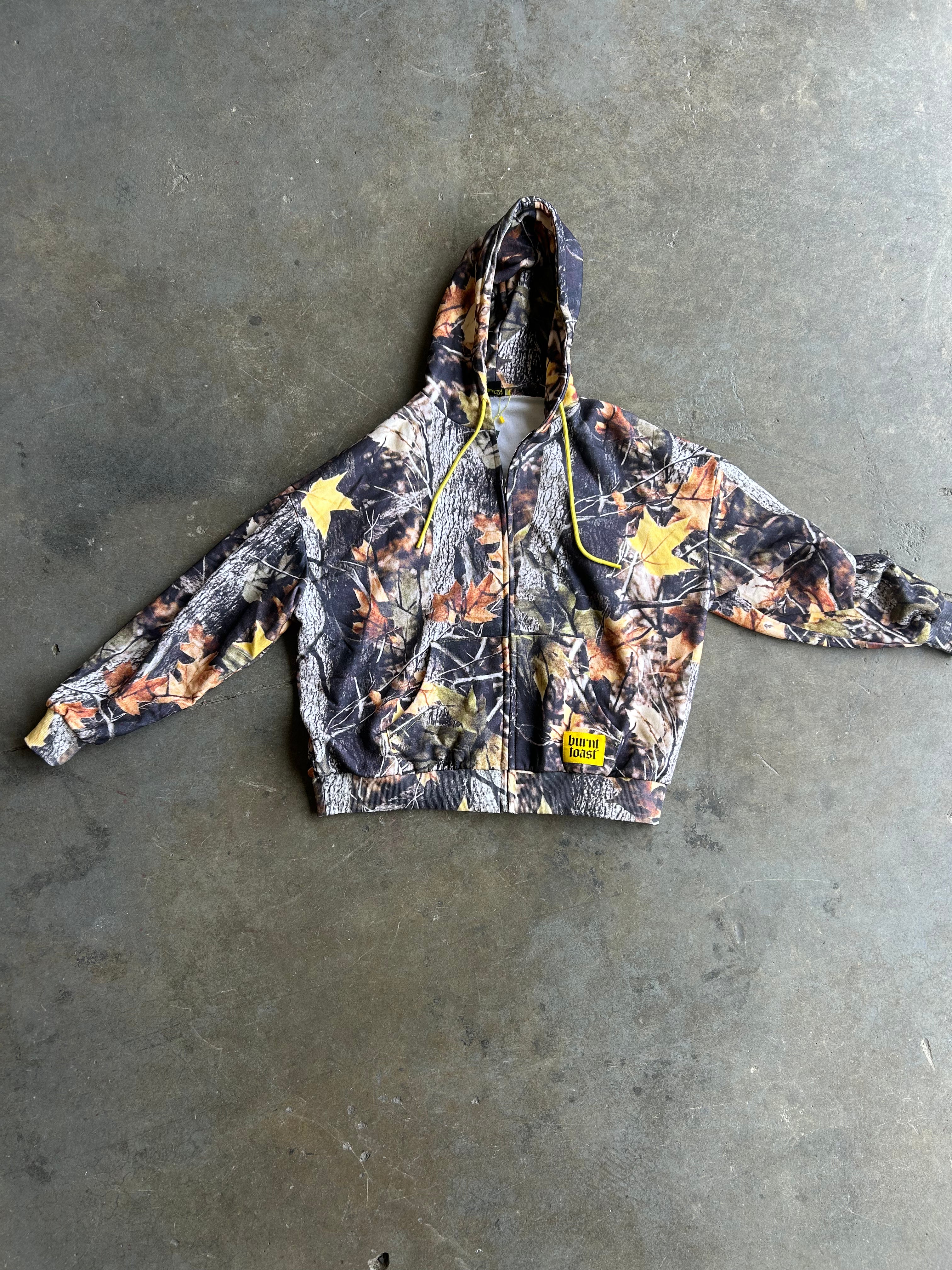 A camouflage zip-up hoodie with yellow stars and a yellow drawstring, laid flat on a gray concrete floor