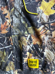 Close-up of a camouflage jacket featuring a woodland leaf and bark print in autumn tones, with a bright yellow drawstring and a yellow Burnt Toast brand patch near the bottom