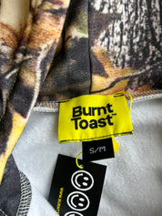 Close-up of a garment's inner neck area showing a yellow Burnt Toast brand label, a black size tag reading S/M, and a black care tag with smiley faces, against a camouflage-patterned fabric background