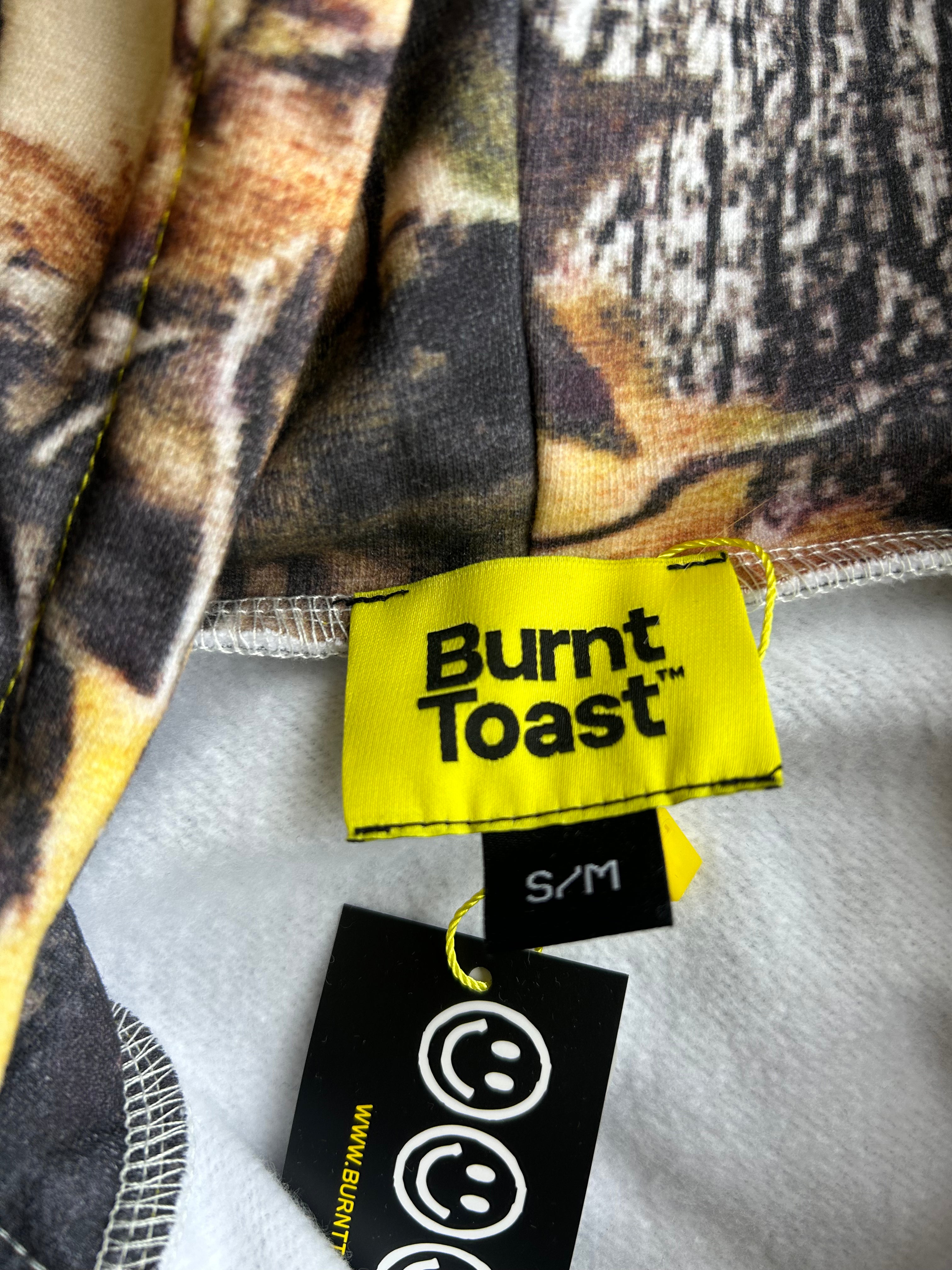 Close-up of a garment's inner neck area showing a yellow Burnt Toast brand label, a black size tag reading S/M, and a black care tag with smiley faces, against a camouflage-patterned fabric background