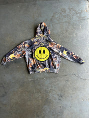 A camouflage hoodie with a large yellow smiley face patch on the chest, laid flat on a gray concrete floor