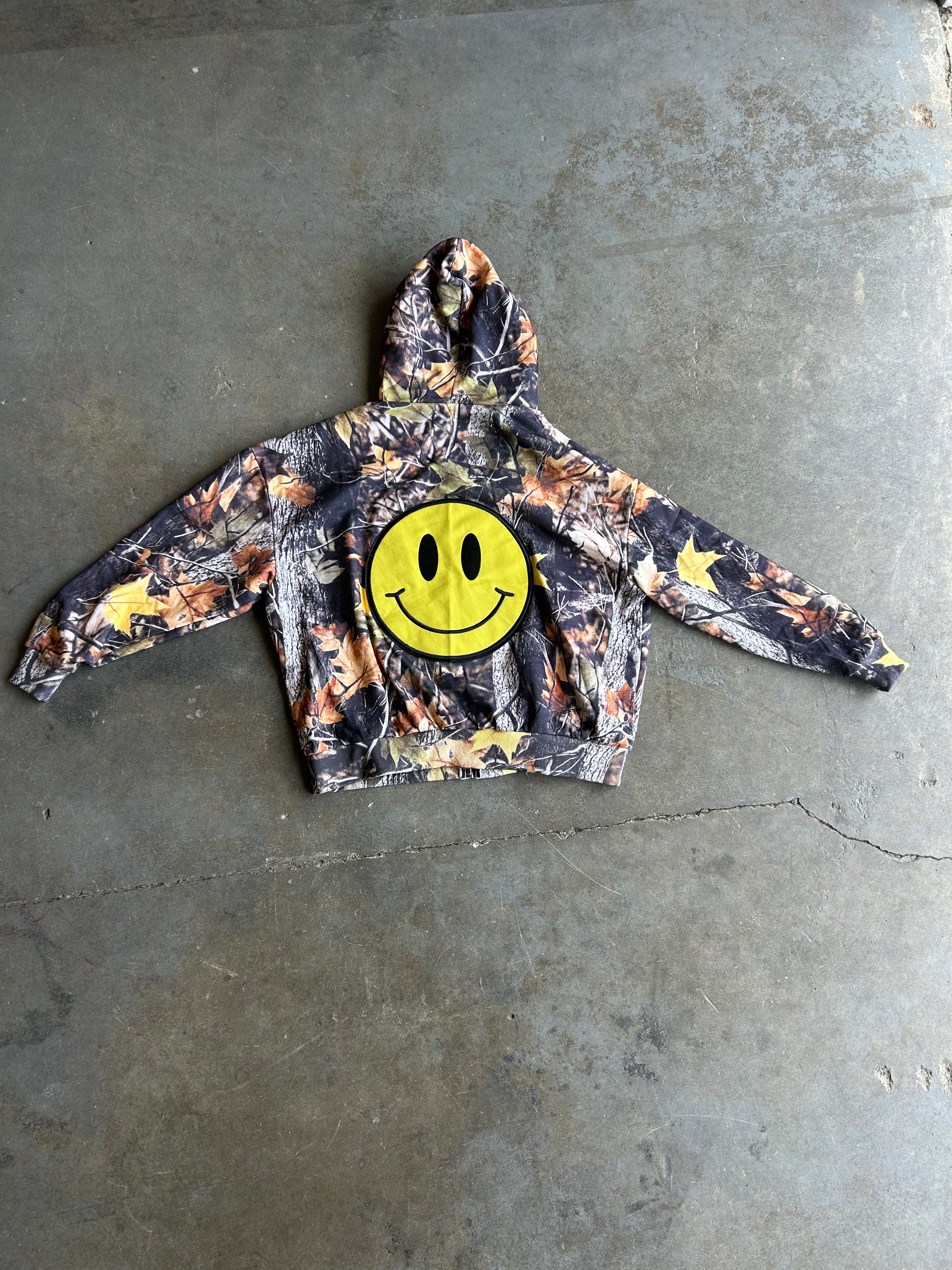 A camouflage hoodie with a large yellow smiley face patch on the chest, laid flat on a gray concrete floor