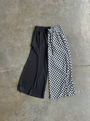 A pair of wide-leg cropped pants laid flat on a concrete floor, featuring a split design with one leg in solid black and the other in a black and white checkerboard pattern