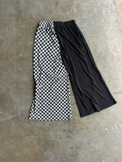 A pair of cropped wide-leg pants laid flat on a concrete floor, featuring one leg in a black and white checkerboard pattern and the other leg in solid black