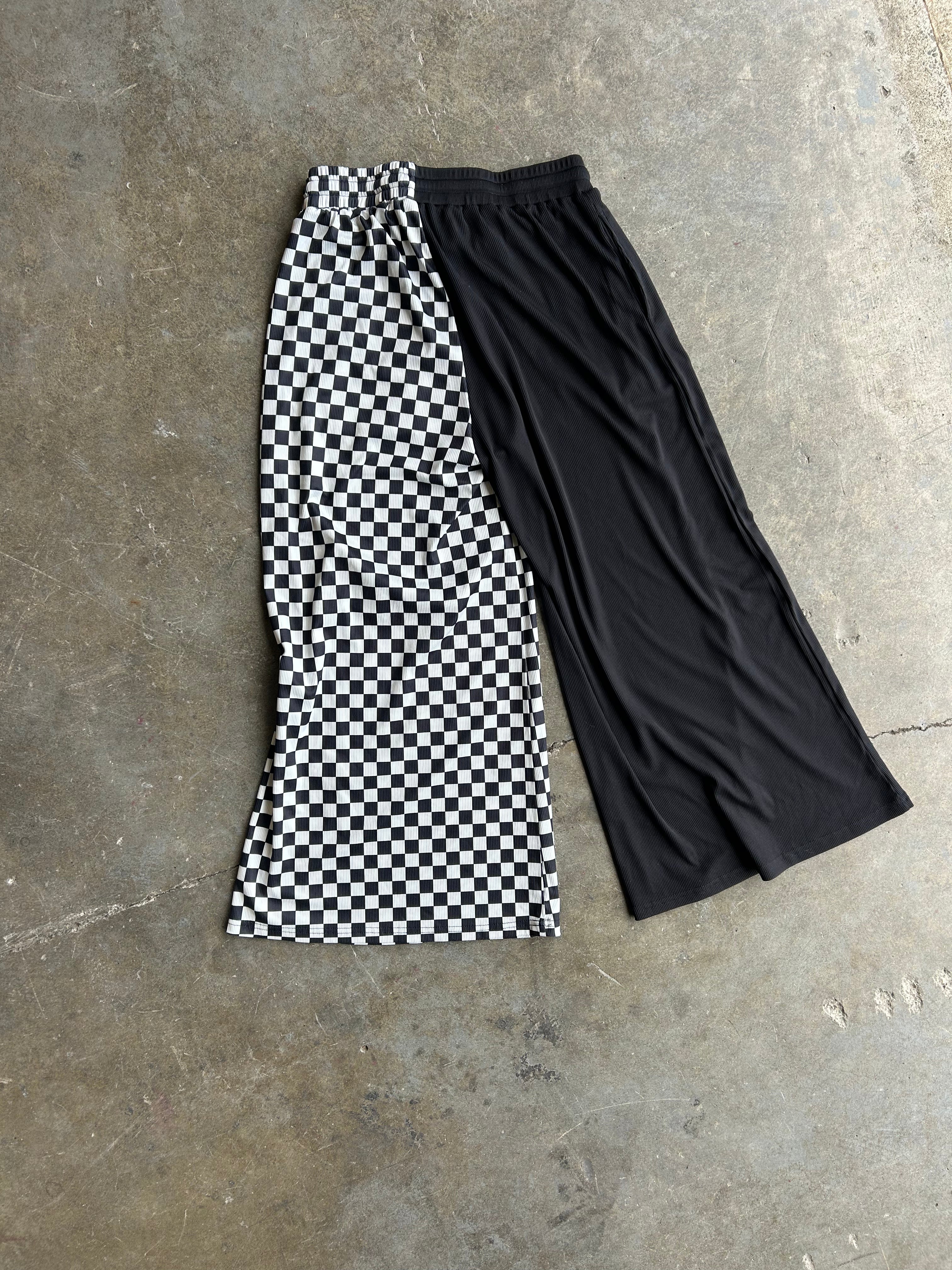 A pair of cropped wide-leg pants laid flat on a concrete floor, featuring one leg in a black and white checkerboard pattern and the other leg in solid black