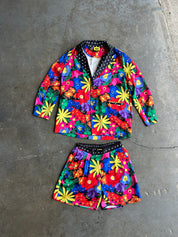 A two-piece children's outfit set consisting of a long-sleeve button-front jacket and matching shorts, both covered in a vibrant multicolor floral print, laid flat on a gray concrete floor