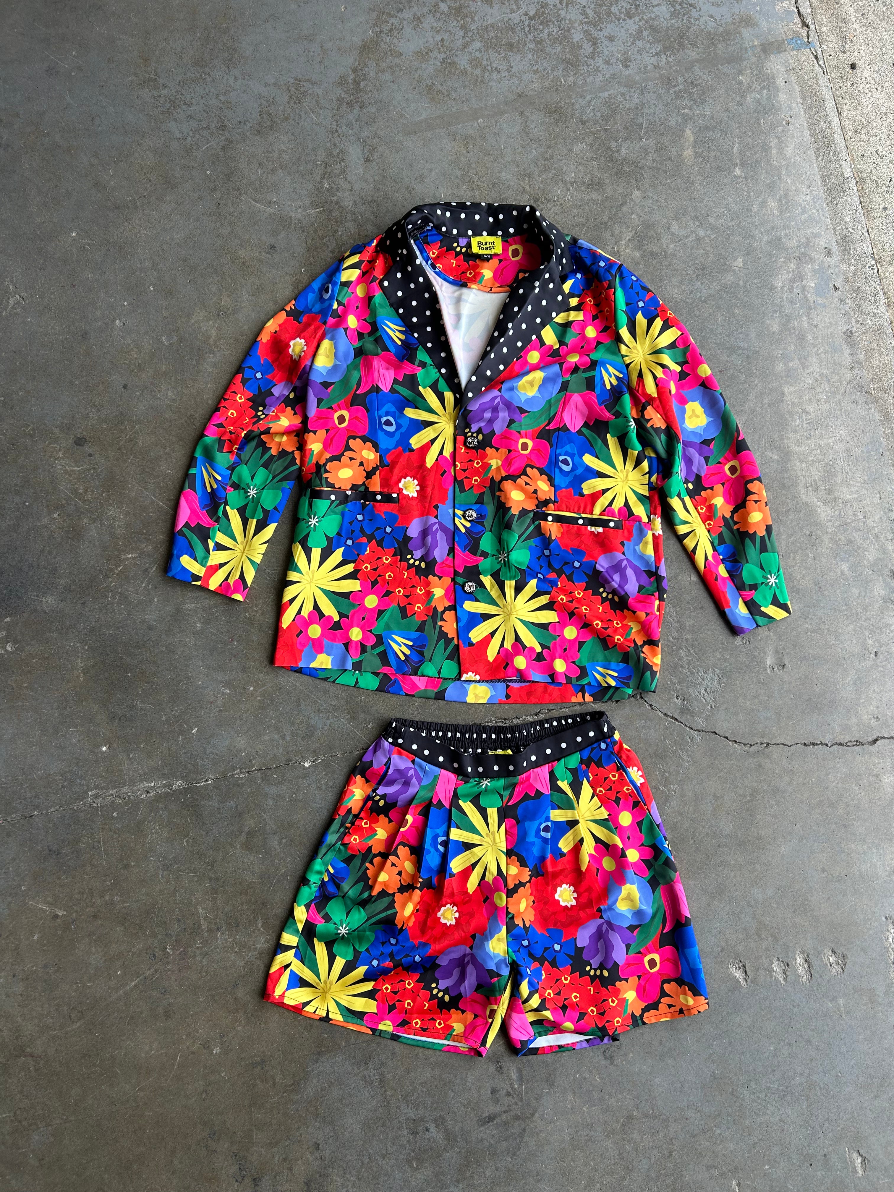 A two-piece children's outfit set consisting of a long-sleeve button-front jacket and matching shorts, both covered in a vibrant multicolor floral print, laid flat on a gray concrete floor