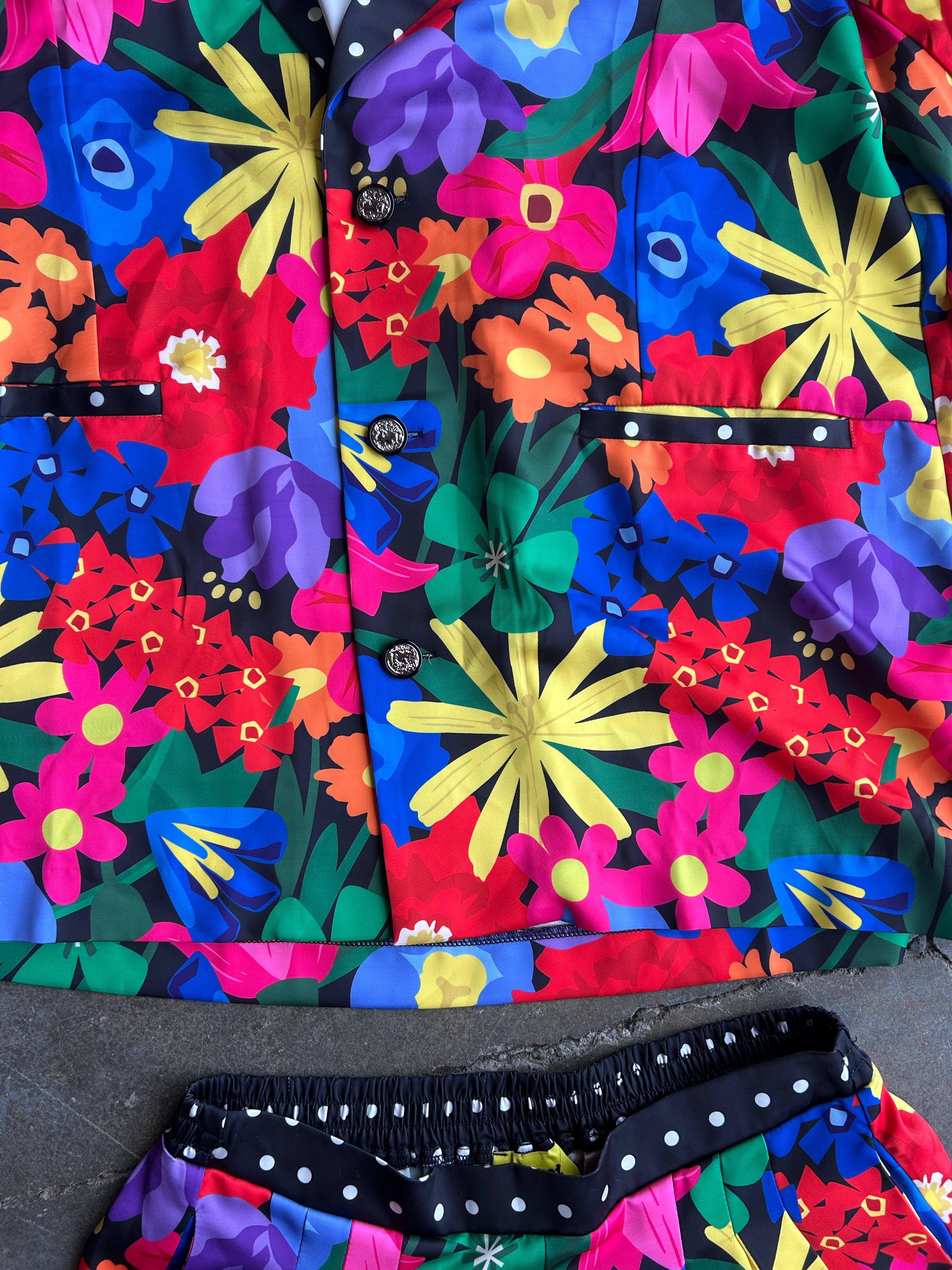 A close-up of a vibrant, multicolor floral print pajama set featuring a button-up shirt and matching shorts. The print contains bold flowers in red, pink, yellow, blue, purple, and green on a black background