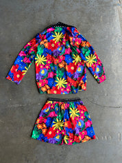 A two-piece girls' outfit set consisting of a long-sleeve crop top and matching shorts, both featuring a vibrant, multicolor floral print with yellow, red, blue, green, and pink flowers on a dark background. The set is laid flat on a textured gray concrete floor
