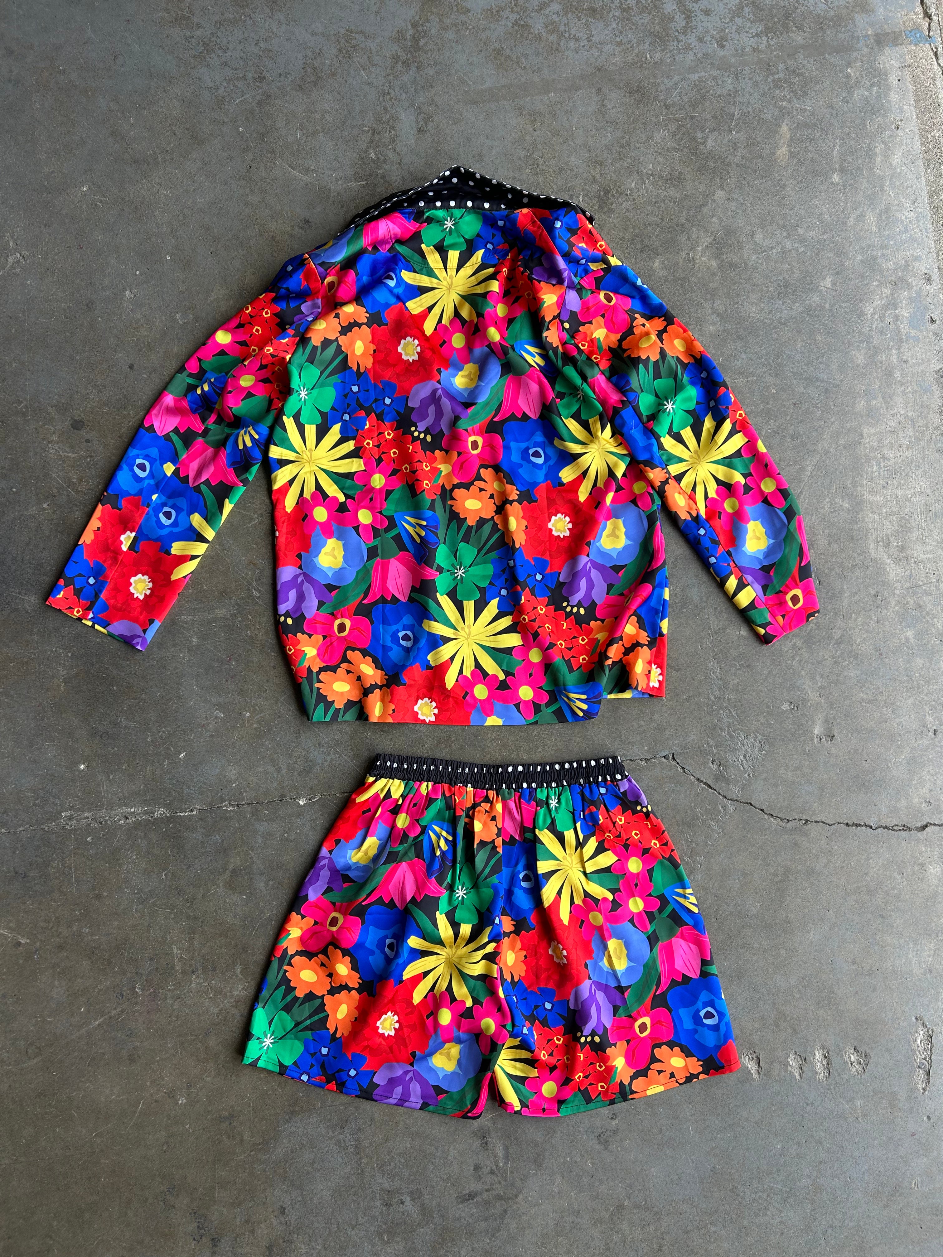 A two-piece girls' outfit set consisting of a long-sleeve crop top and matching shorts, both featuring a vibrant, multicolor floral print with yellow, red, blue, green, and pink flowers on a dark background. The set is laid flat on a textured gray concrete floor