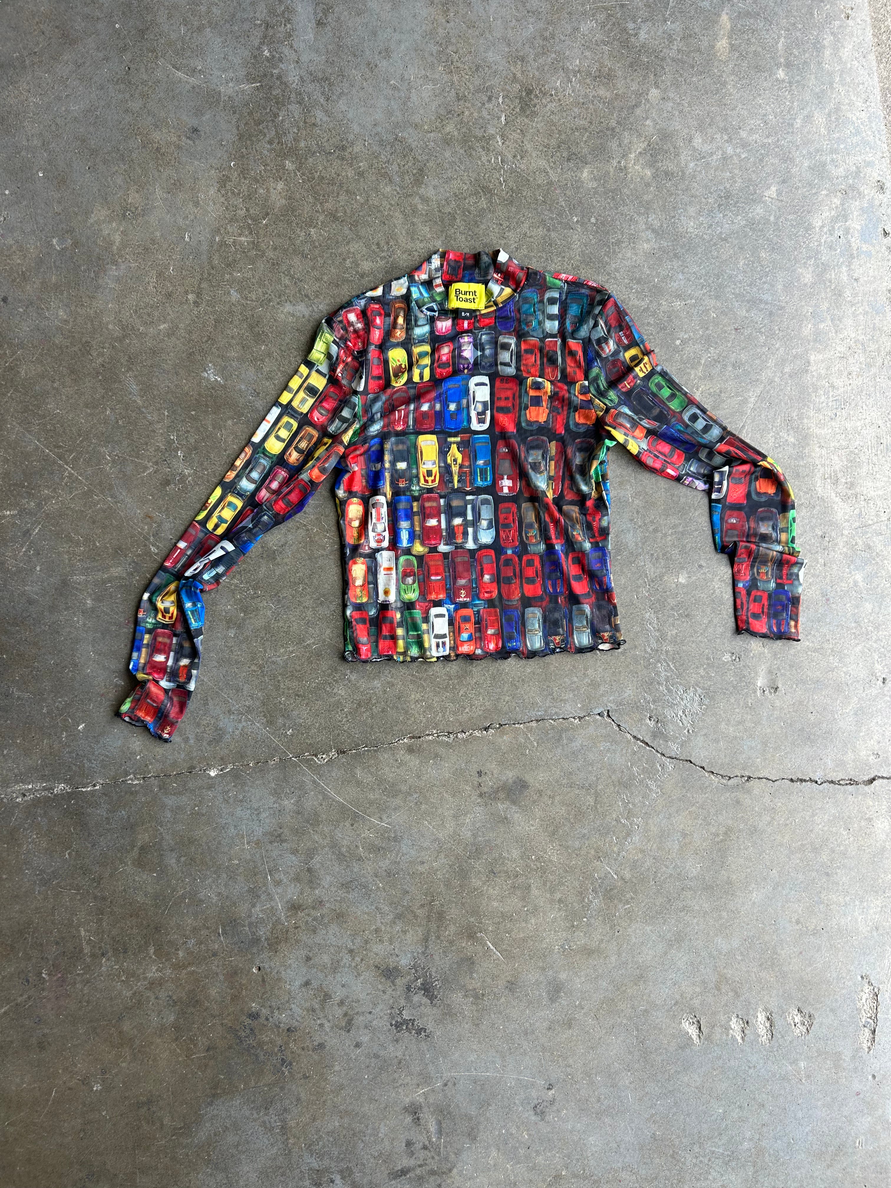 A long-sleeve crewneck shirt with a dense, colorful print of classic cars in red, blue, yellow, and black, laid flat on a textured gray concrete floor
