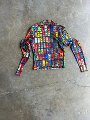 A long-sleeve crop top with a colorful busy pattern of miniature cars, laid flat on a textured gray concrete floor