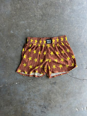 A pair of brown shorts with a yellow star pattern and vertical stripes, laid flat on a gray concrete floor. A black brand label reading 'BURNT TOAST' is visible at the waistband