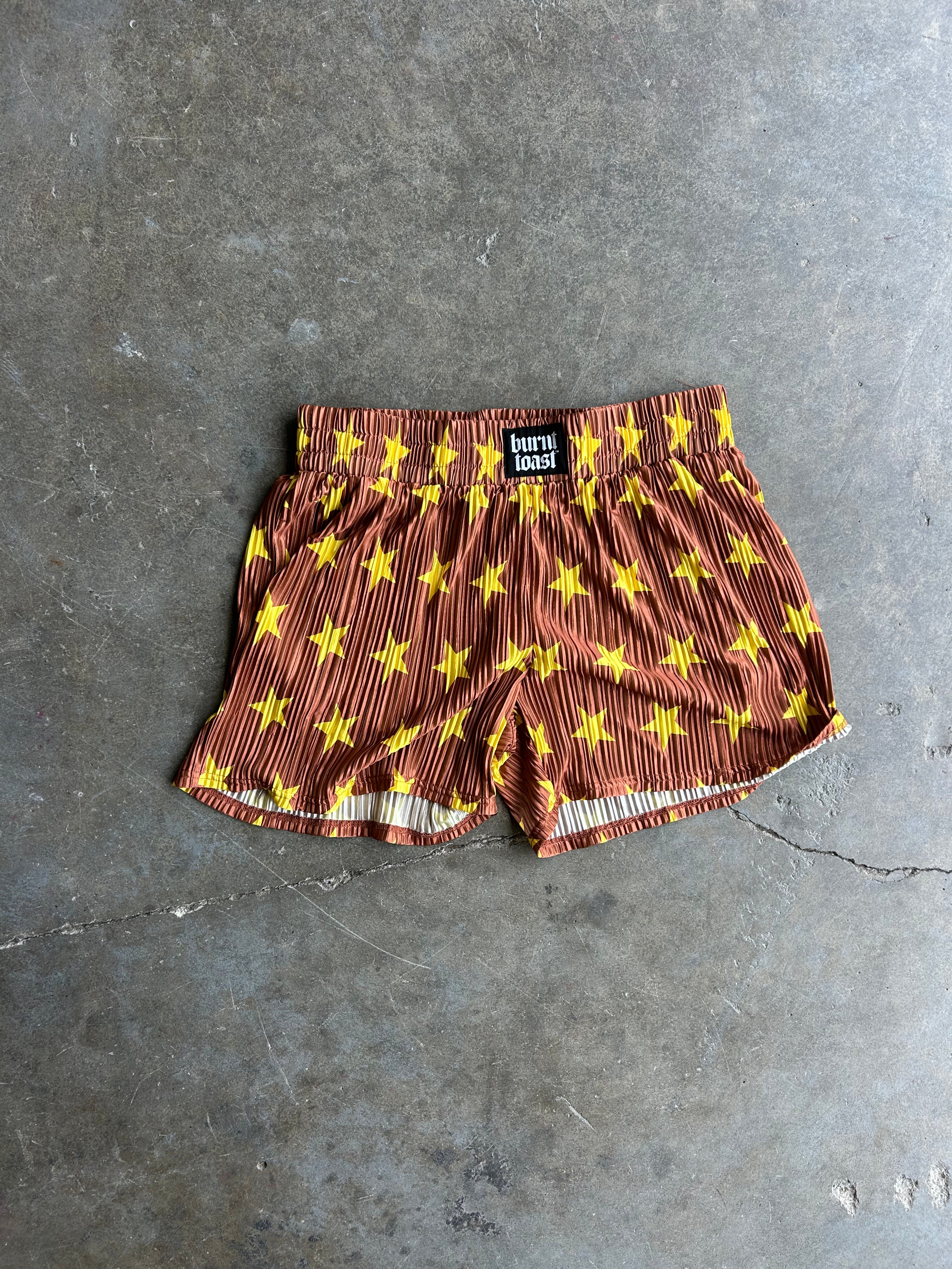 A pair of brown shorts with a yellow star pattern and vertical stripes, laid flat on a gray concrete floor. A black brand label reading 'BURNT TOAST' is visible at the waistband