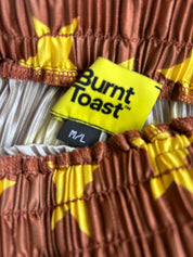 Close-up of a garment label reading 'Burnt Toast' with 'M/L' below it, showing part of a pleated fabric in rust brown and yellow with white pleated trim