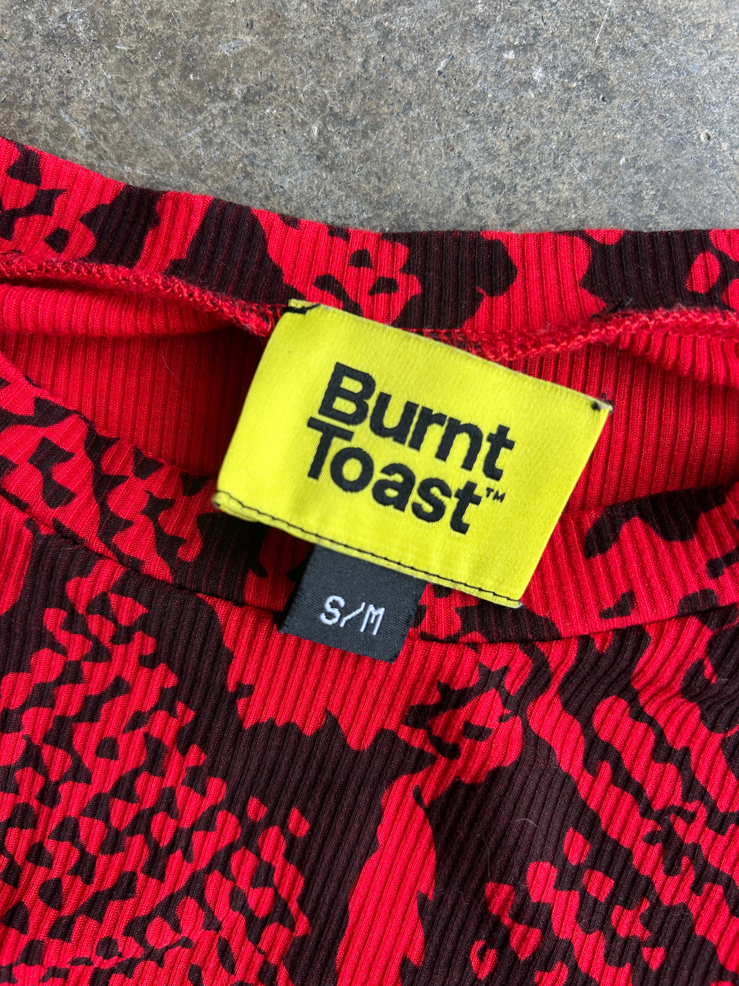 Close-up of a ribbed red and black patterned garment with a bright yellow Burnt Toast brand label and a small black size tag reading S/M
