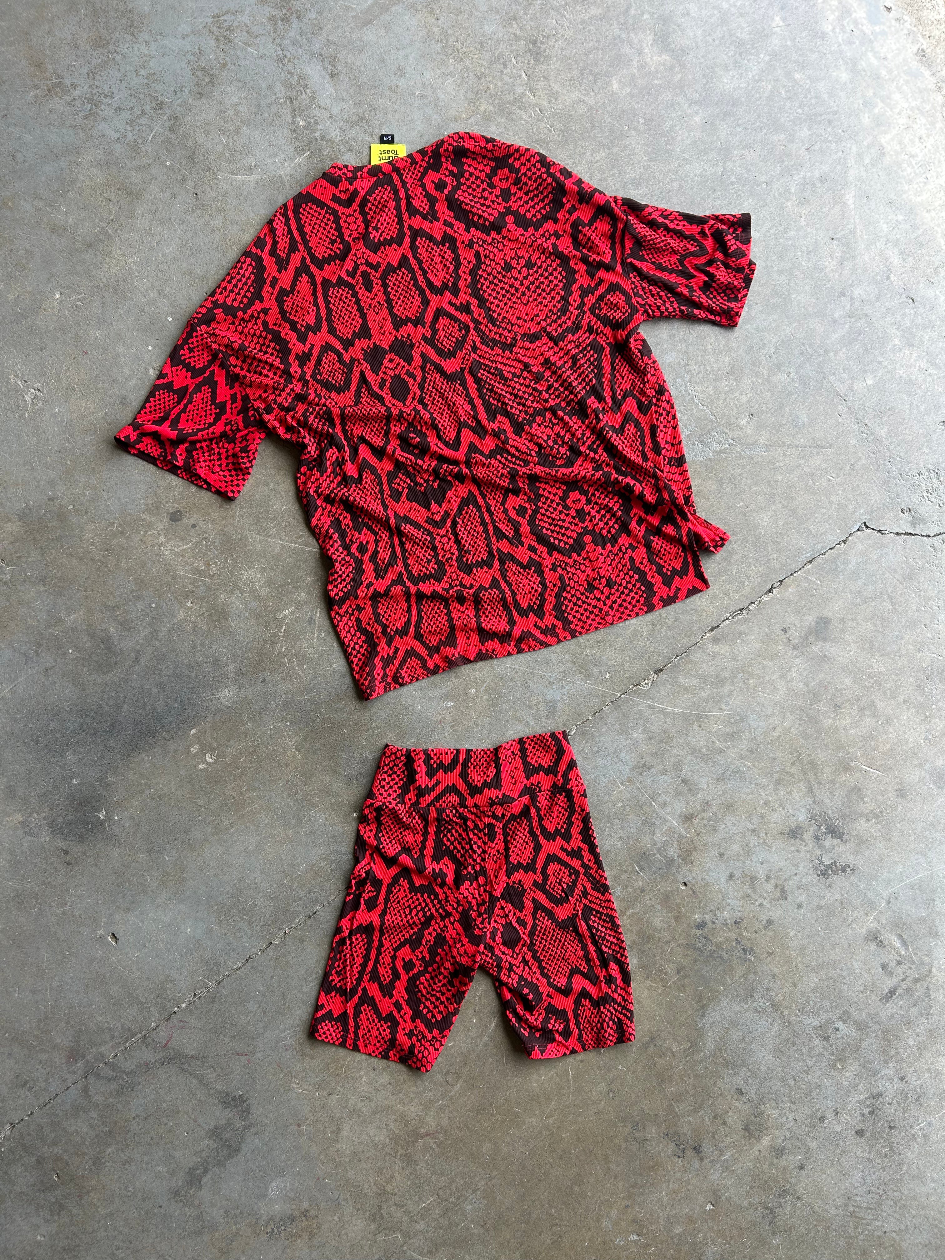 A red and black snakeskin print matching set consisting of a short-sleeve top and shorts, laid flat on a gray concrete floor