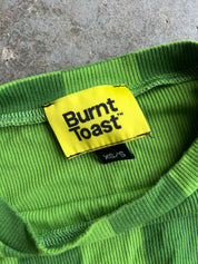 Close-up of a bright green ribbed knit garment showing the inner neck label, which reads 'Burnt Toast' in black text on a yellow patch, with a small black size tag below reading 'XS/S'