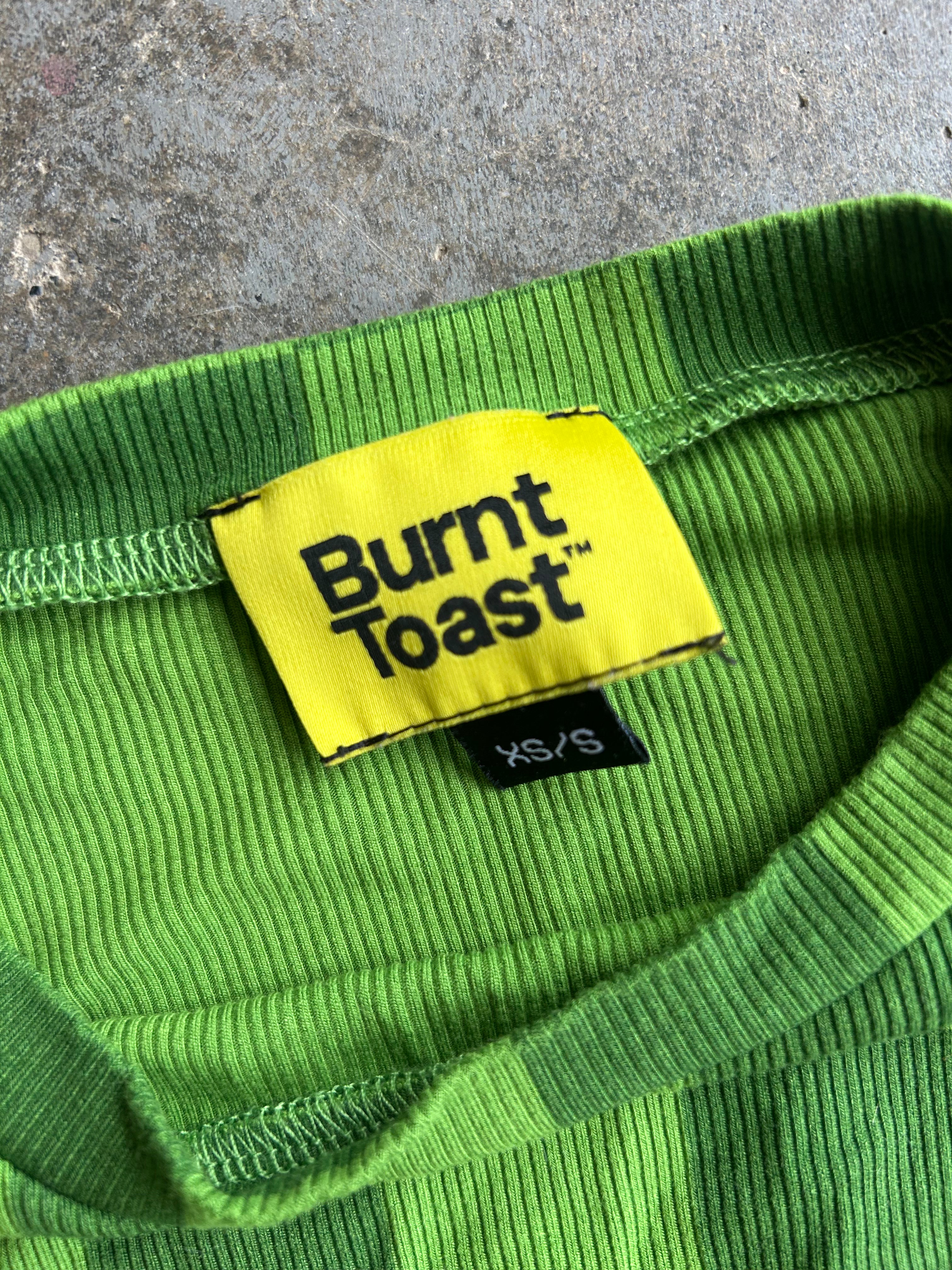Close-up of a bright green ribbed knit garment showing the inner neck label, which reads 'Burnt Toast' in black text on a yellow patch, with a small black size tag below reading 'XS/S'