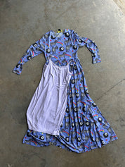 A long-sleeve dress laid flat on a concrete floor, featuring a blue base with a black, white, and multicolor zebra-and-eye print on one side, and a solid light lavender panel on the other