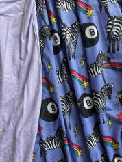 A close-up of a light blue fabric with a playful print of black-and-white zebras, black 8-ball game pieces, rainbow accents, and yellow stars, next to a plain lavender fabric