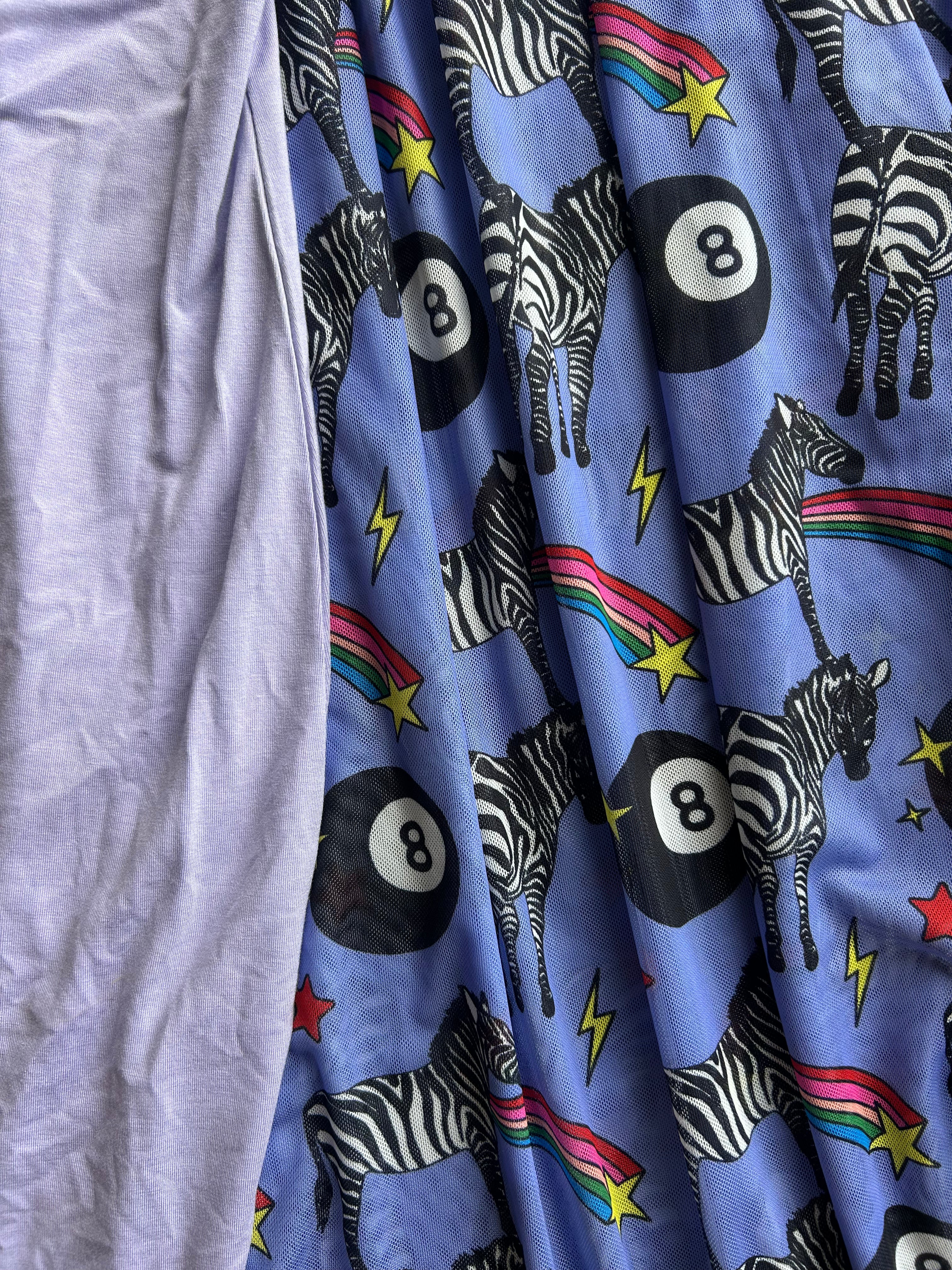 A close-up of a light blue fabric with a playful print of black-and-white zebras, black 8-ball game pieces, rainbow accents, and yellow stars, next to a plain lavender fabric