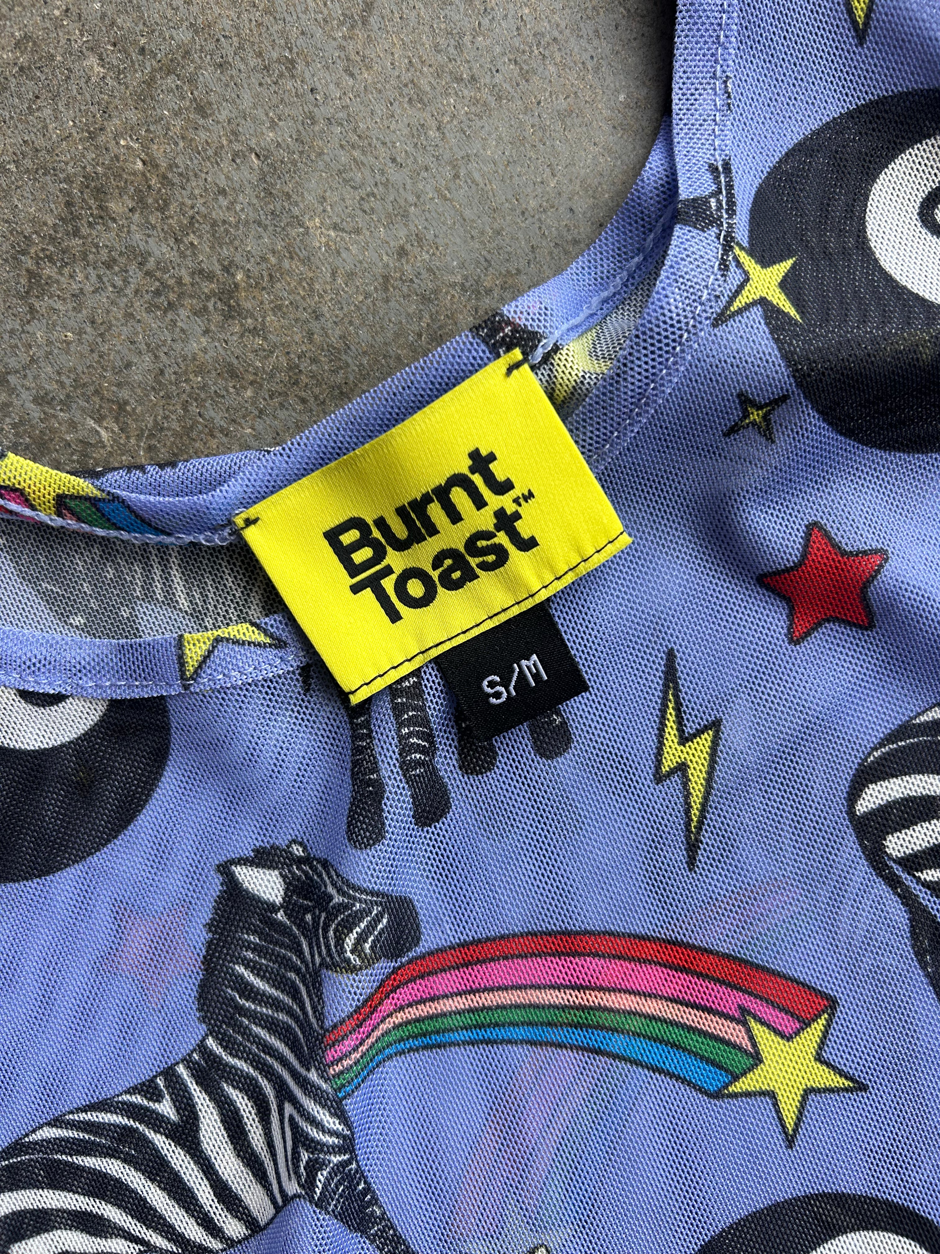 Close-up of a Burnt Toast garment with a bright yellow brand label and a colorful zebra-and-rainbow print on a light blue-purple background