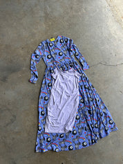 A long-sleeved maxi dress with a blue, black, and red psychedelic print, laid flat on a concrete floor with a light purple undergarment draped over the front
