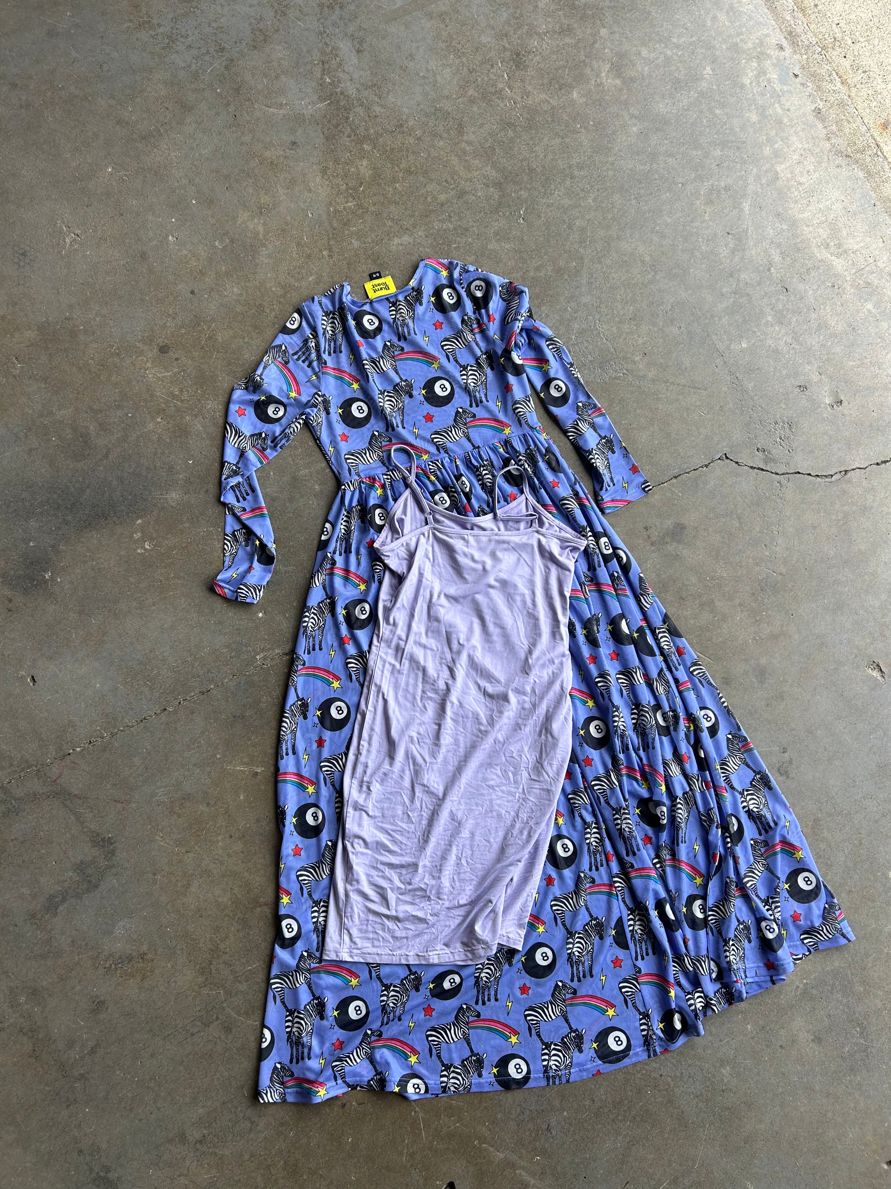 A long-sleeved maxi dress with a blue, black, and red psychedelic print, laid flat on a concrete floor with a light purple undergarment draped over the front