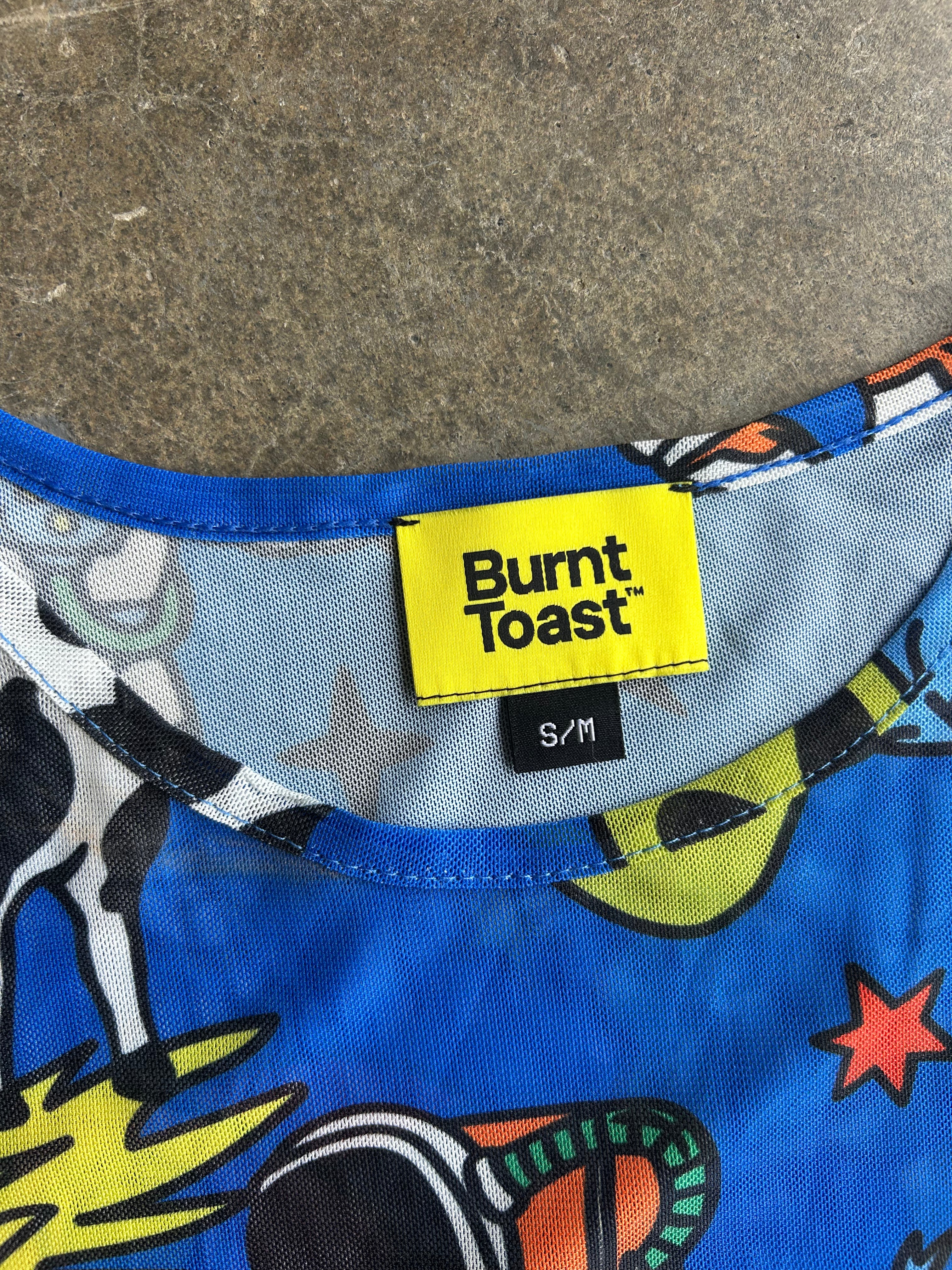 Close-up of a Burnt Toast t-shirt collar showing a bright yellow brand label with 'Burnt Toast' printed in black, alongside a black size tag reading 'S/M'. The shirt features a colorful cartoon space-themed print on a blue base, set against a gray concrete floor background