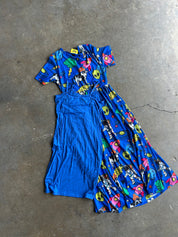 A blue dress with a split design, featuring a solid blue left side and a right side with a colorful cartoon print on a blue background, laid flat on a concrete floor