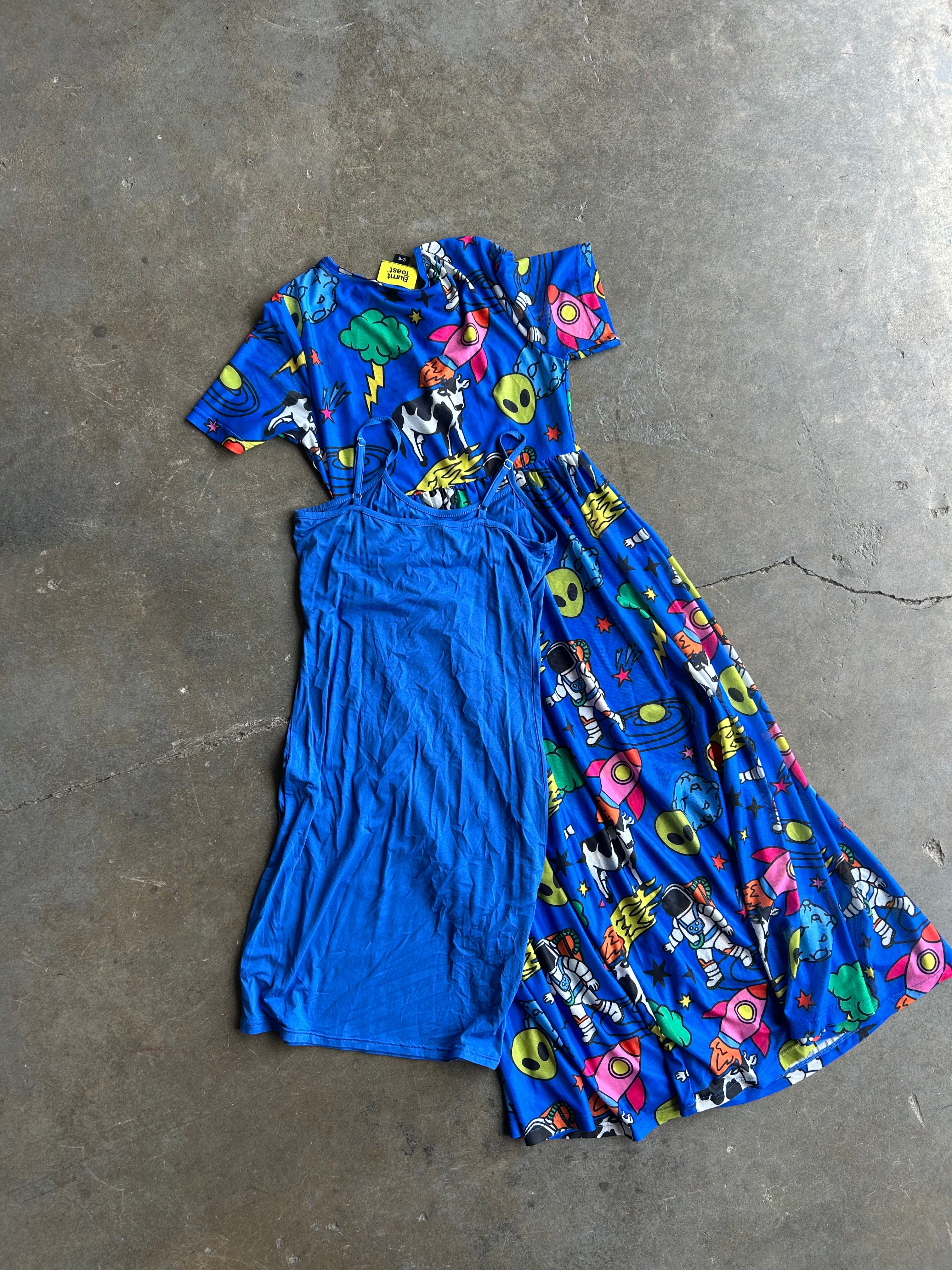 A blue dress with a split design, featuring a solid blue left side and a right side with a colorful cartoon print on a blue background, laid flat on a concrete floor
