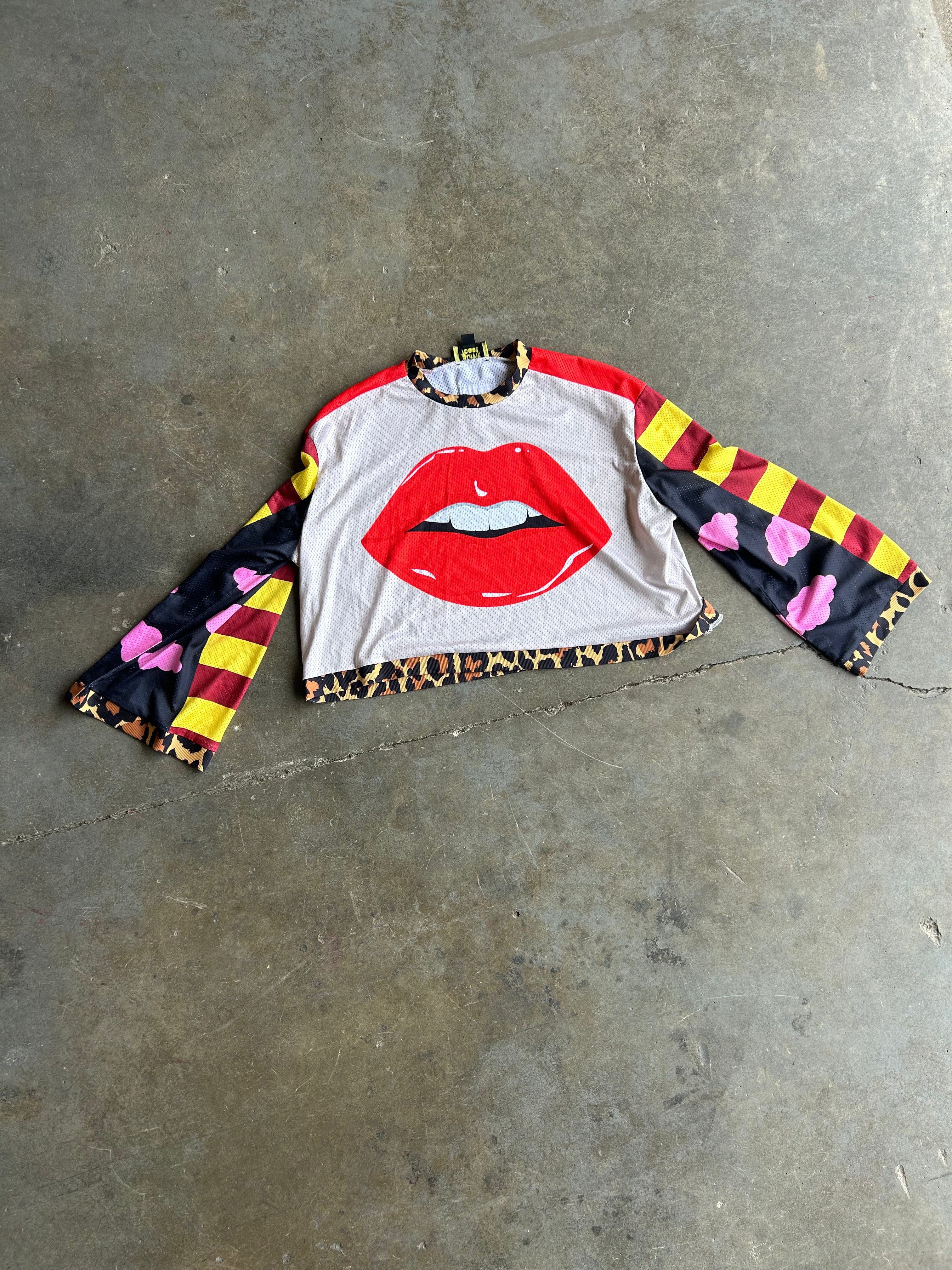 A long-sleeve crop top with a white body, a large red lips graphic on the front, red and yellow striped sleeves with pink floral accents, and leopard print trim, laid flat on a gray concrete floor