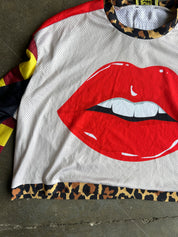A white mesh long-sleeve shirt with a large red lips graphic on the front, laid flat on a concrete floor. The shirt features a leopard print hem, leopard print inner collar, and multicolor patterned sleeves