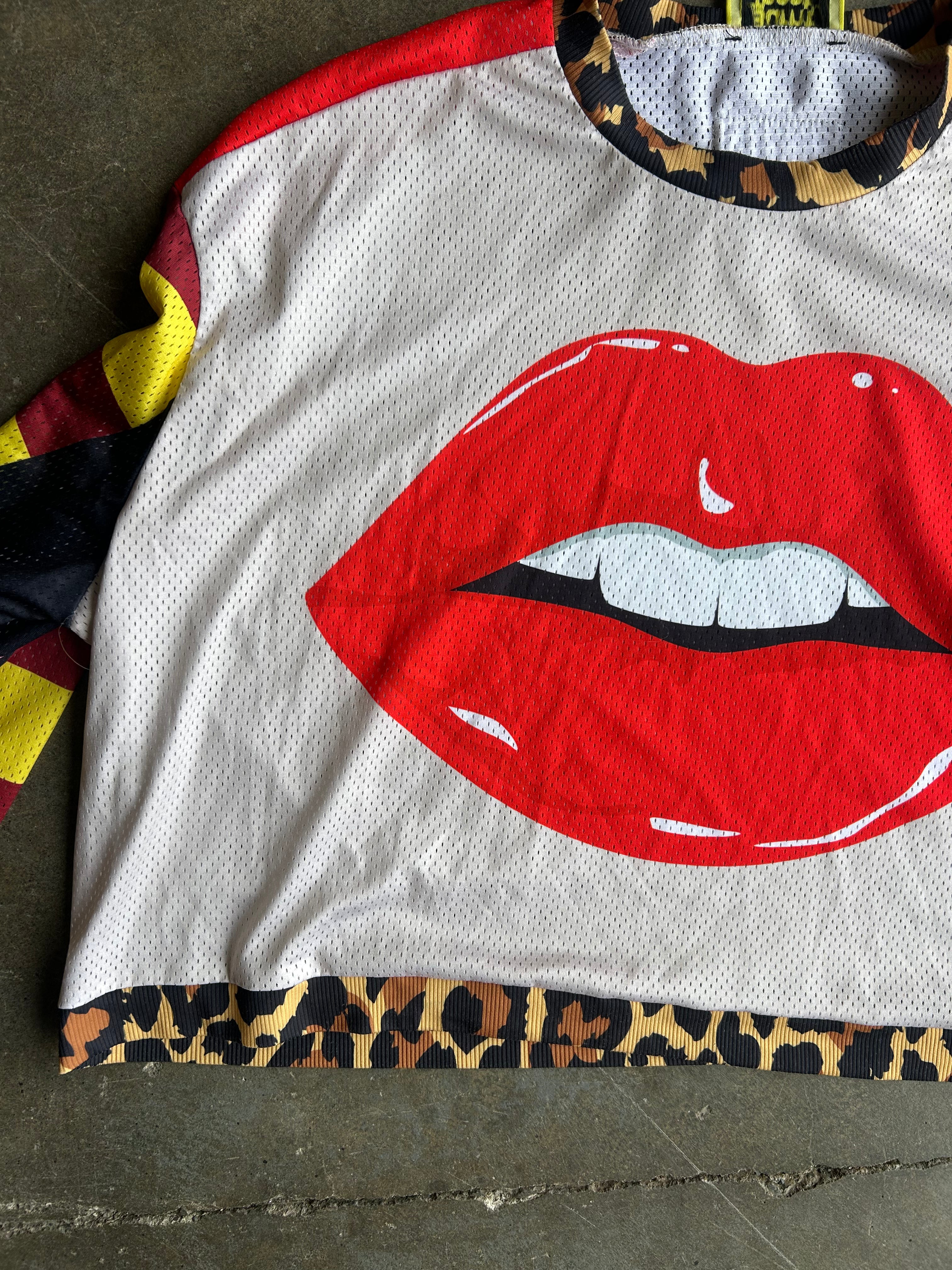 A white mesh long-sleeve shirt with a large red lips graphic on the front, laid flat on a concrete floor. The shirt features a leopard print hem, leopard print inner collar, and multicolor patterned sleeves