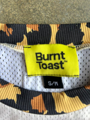Close-up of a Burnt Toast garment neck label on a white mesh collar with a black, orange, and yellow abstract pattern around the ribbed trim