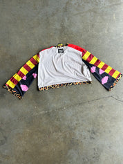A cropped long-sleeve shirt laid flat on a concrete floor, featuring a white body with red and yellow striped sleeves, pink cloud motifs on the sleeves, and a leopard-print hem