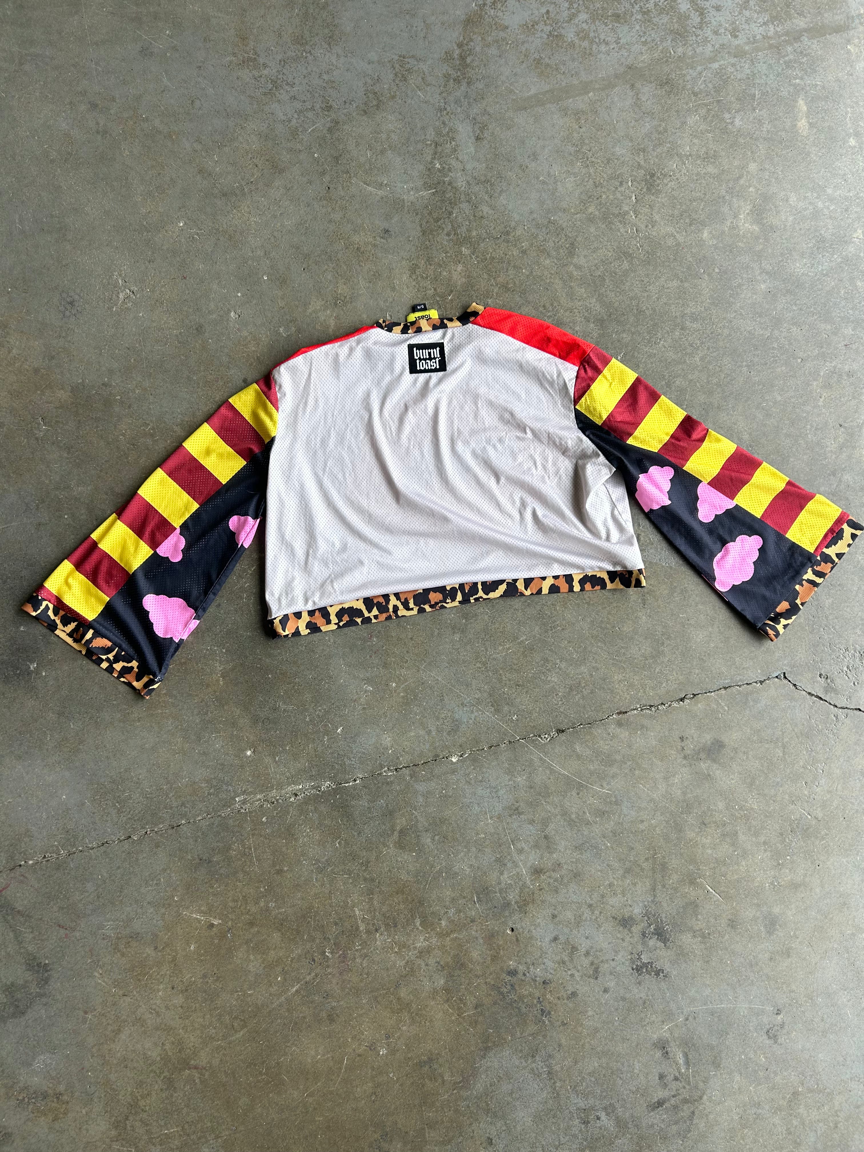 A cropped long-sleeve shirt laid flat on a concrete floor, featuring a white body with red and yellow striped sleeves, pink cloud motifs on the sleeves, and a leopard-print hem
