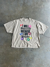 A beige graphic t-shirt laid flat on a gray concrete floor. The shirt features a bold text graphic reading 'I WILL Succeed BECAUSE I AM Insane' in black, surrounded by colorful billiard balls in red, pink, blue, purple, green, and yellow