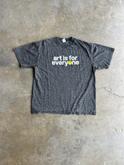 A dark gray leopard-print t-shirt with the text 'art is for everyone' printed in white, featuring a small yellow smiley face on the letter 'o', laid flat on a concrete floor