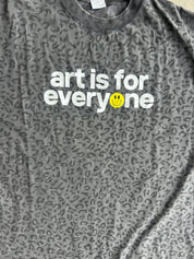 Close up of a dark gray leopard-print t-shirt with the text 'art is for everyone' printed in white, featuring a small yellow smiley face on the letter 'o', laid flat on a concrete floor
