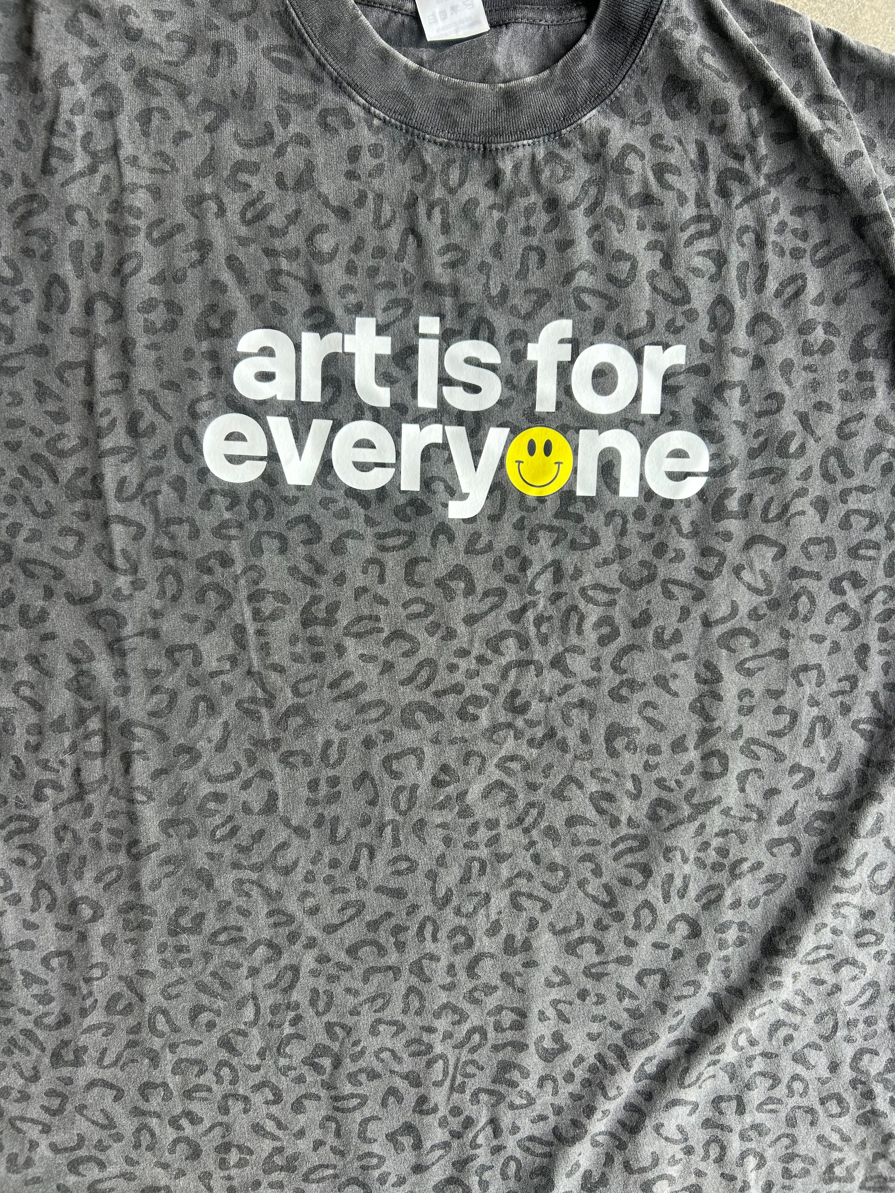 Close up of a dark gray leopard-print t-shirt with the text 'art is for everyone' printed in white, featuring a small yellow smiley face on the letter 'o', laid flat on a concrete floor