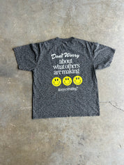 A dark gray short-sleeve t-shirt with a subtle leopard print pattern, featuring a white graphic text that reads 'Don't Worry about what others are making' above three yellow smiley faces and the phrase 'keep creating!' at the bottom. The shirt is laid flat on a textured concrete floor
