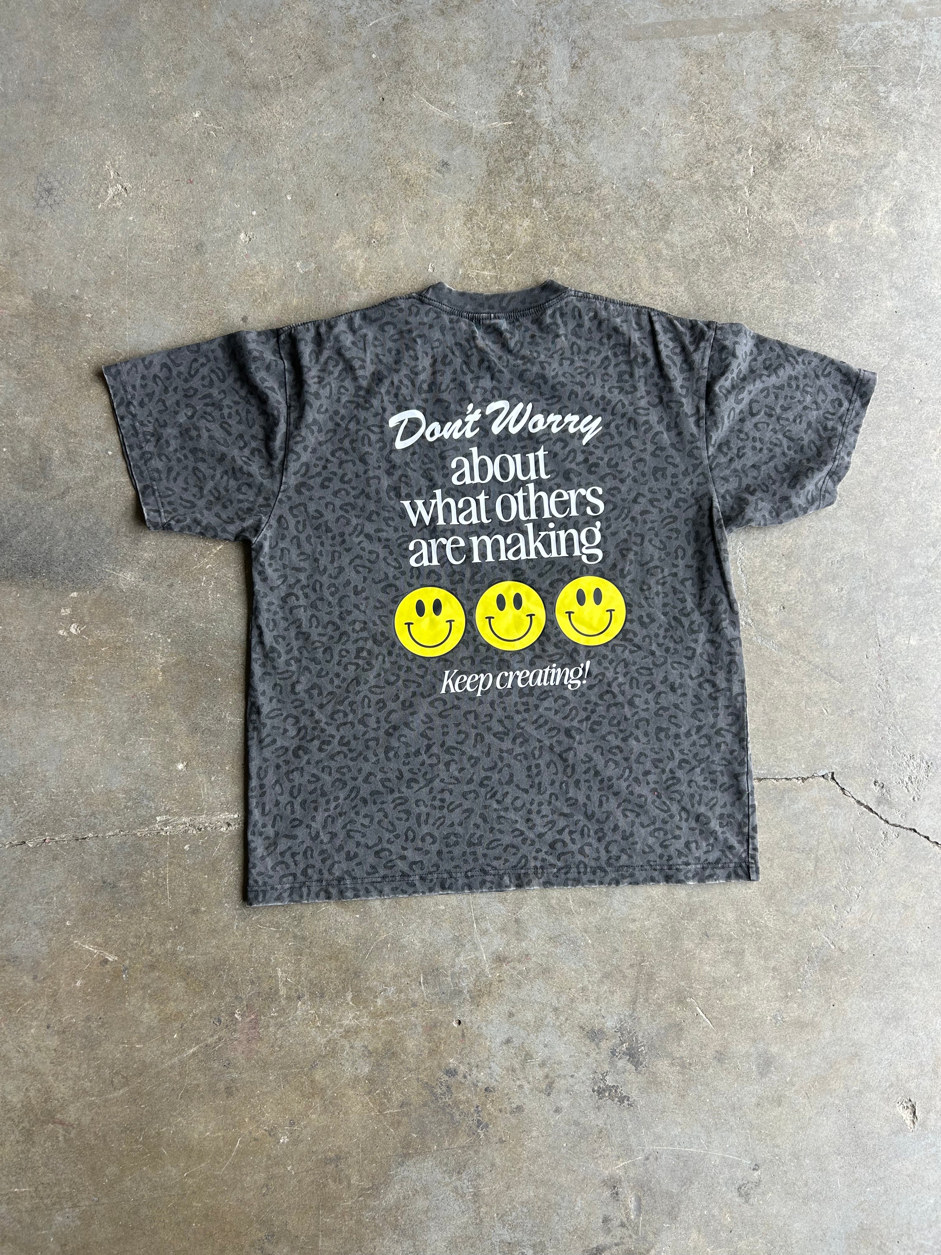 A dark gray short-sleeve t-shirt with a subtle leopard print pattern, featuring a white graphic text that reads 'Don't Worry about what others are making' above three yellow smiley faces and the phrase 'keep creating!' at the bottom. The shirt is laid flat on a textured concrete floor