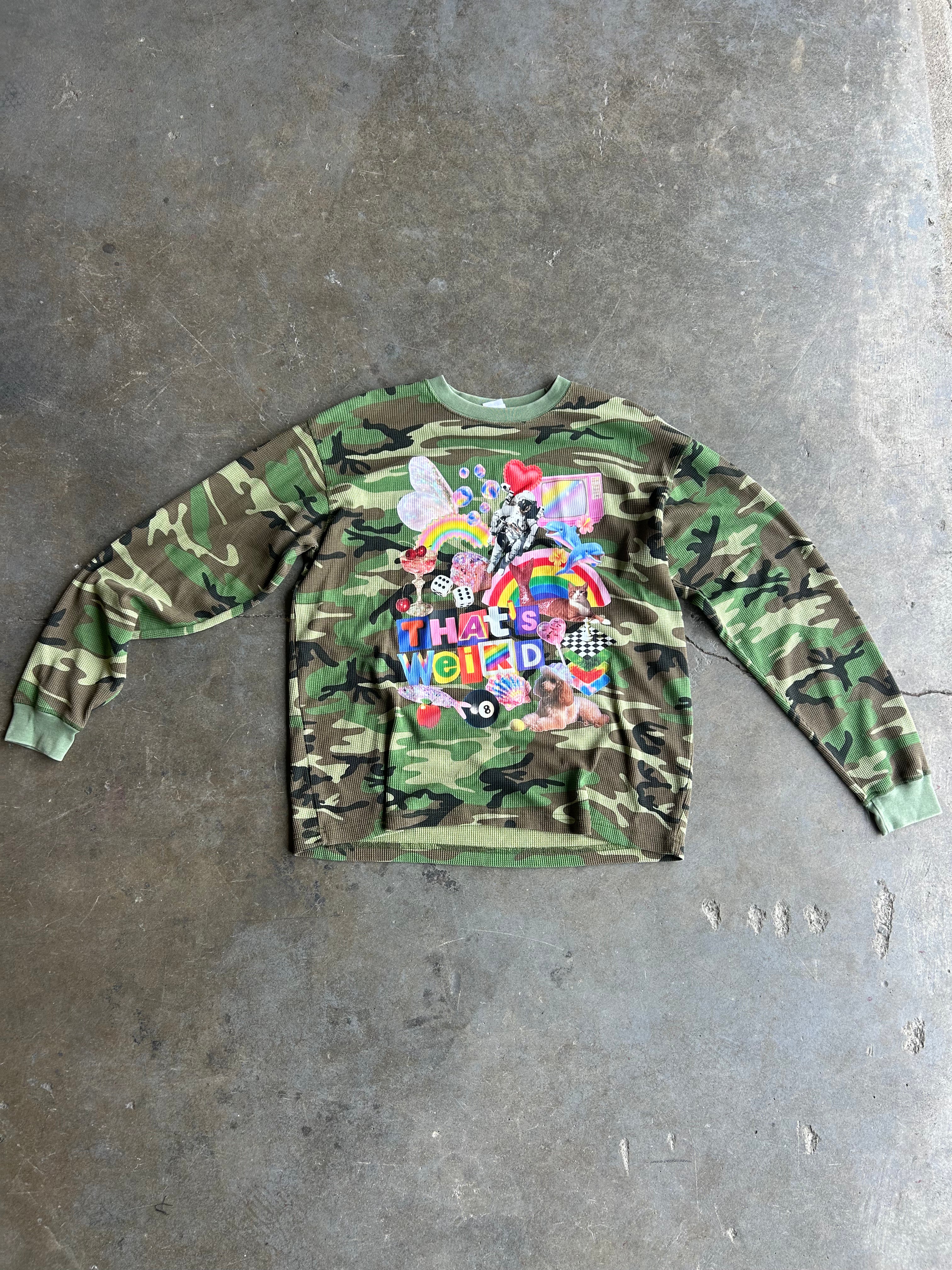 Long sleeve camo waffle tee with a collage on the front with the text That's Weird, flat lay frontal view on a concrete surface