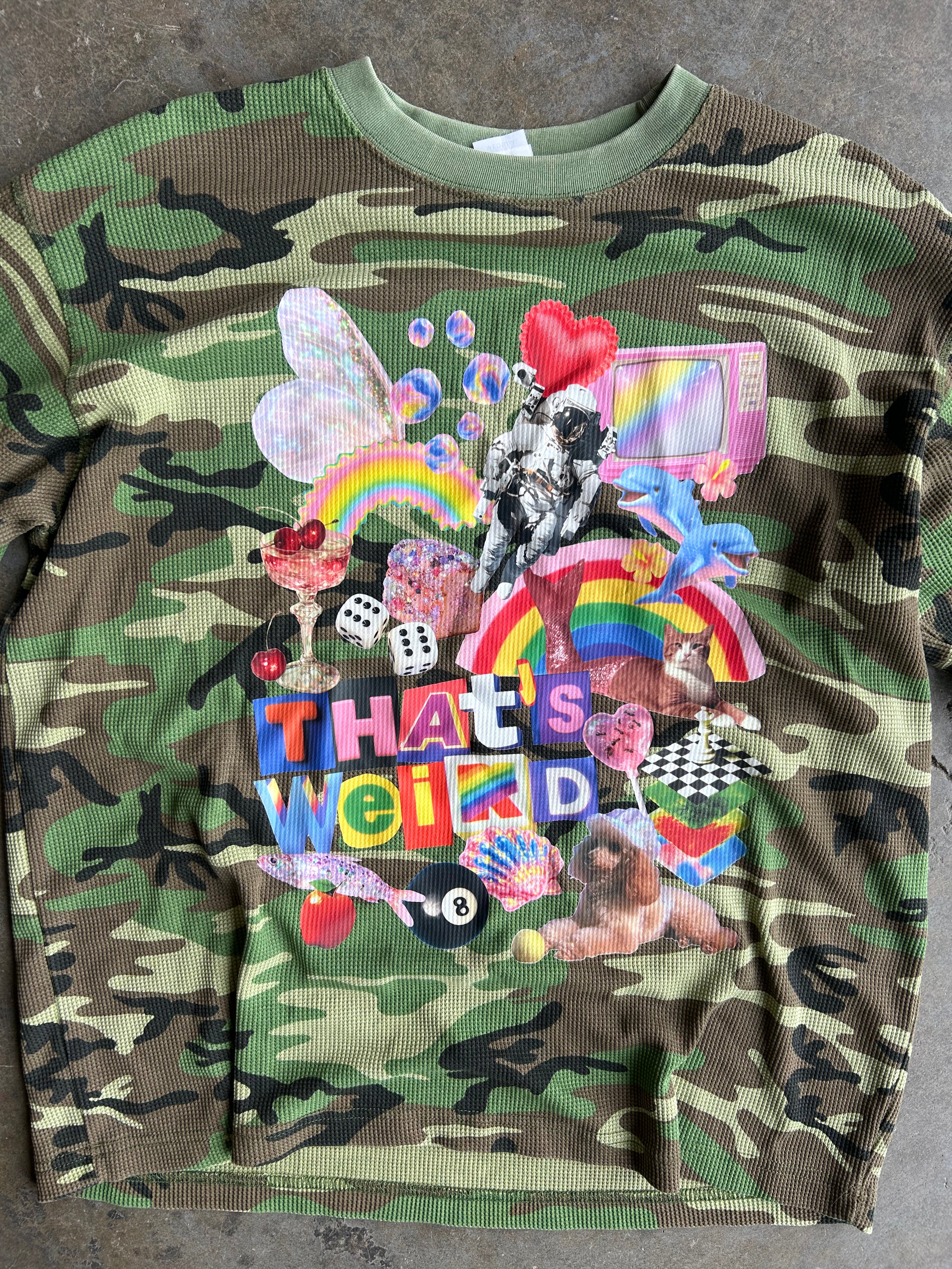 Close up of a long sleeve camo waffle tee with a collage on the front with the text That's Weird, flat lay frontal view on a concrete surface