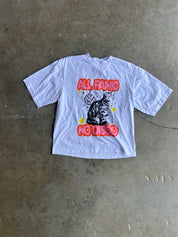 A white short-sleeve graphic t-shirt laid flat on a concrete floor. The shirt features a black cat illustration with red bubble text reading 'ALL PANIC NO DISCO' and small yellow stars