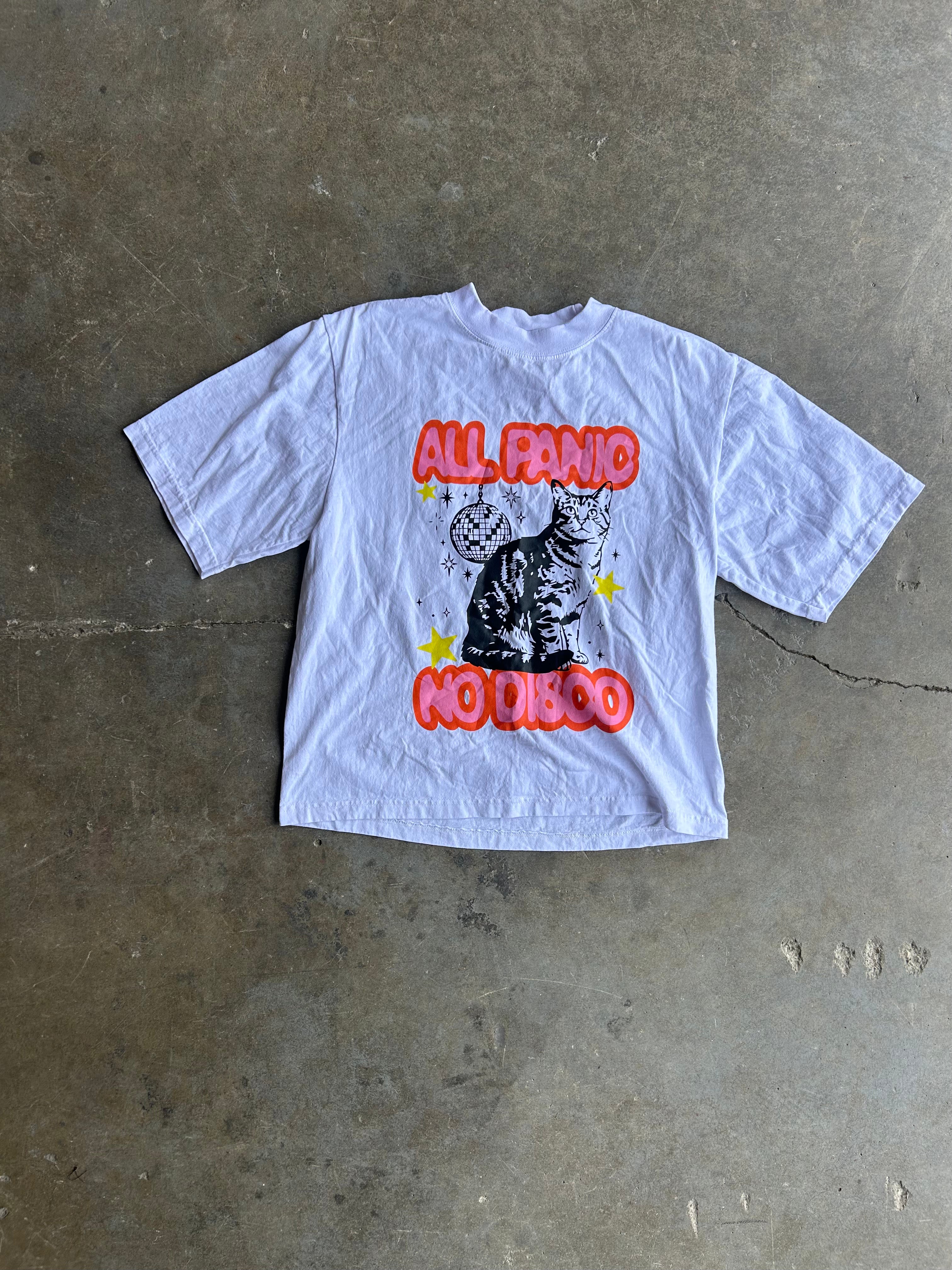 A white short-sleeve graphic t-shirt laid flat on a concrete floor. The shirt features a black cat illustration with red bubble text reading 'ALL PANIC NO DISCO' and small yellow stars