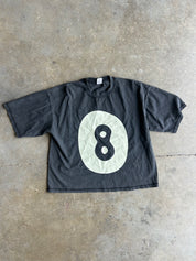A faded black short-sleeve t-shirt with a large white circle graphic on the front featuring a black number 8, laid flat on a concrete floor