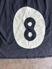 A close-up of a dark black fabric shirt, featuring a large cream-colored oval with a black number 8 printed inside it
